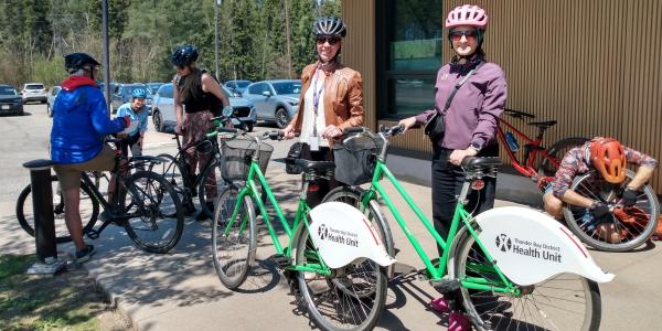 two women with helmets and sunglasses stand beside TBDHU branded bicycles